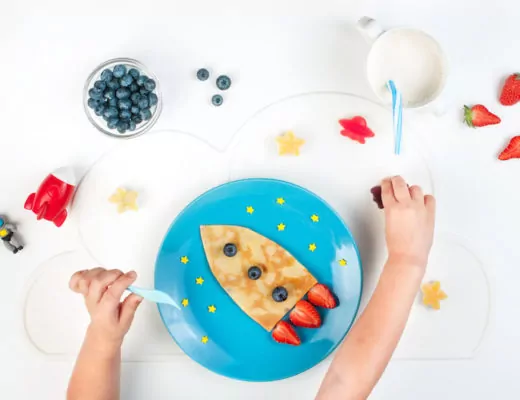 Kinderhände von oben vor einem Teller mit Pfannkuchen in Form einer Rakete