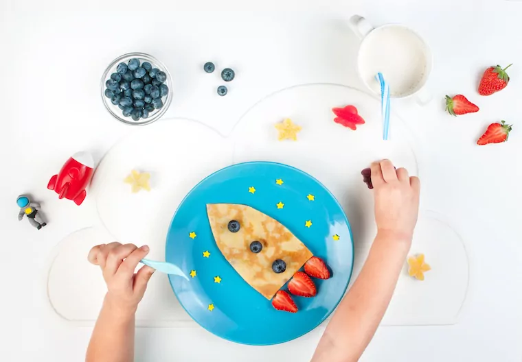 Kinderhände von oben vor einem Teller mit Pfannkuchen in Form einer Rakete