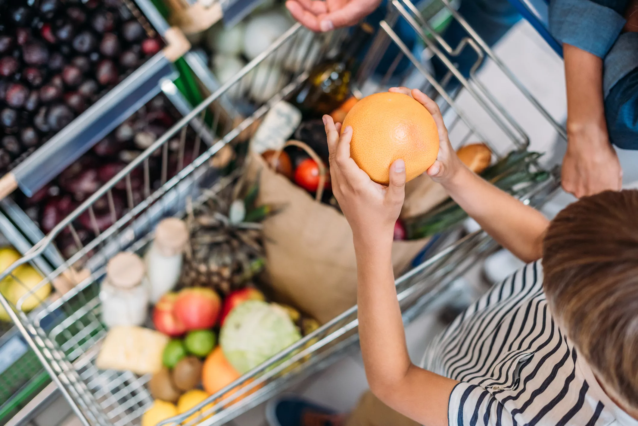 Kinderhände, die eine Grapefruit in einen gefüllten Einkaufwagen legen