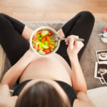 Schwangere Frau isst eine Salat-Bowl auf dem Sofa
