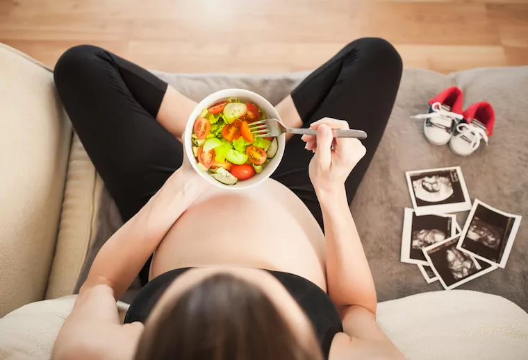 Schwangere Frau isst eine Salat-Bowl auf dem Sofa