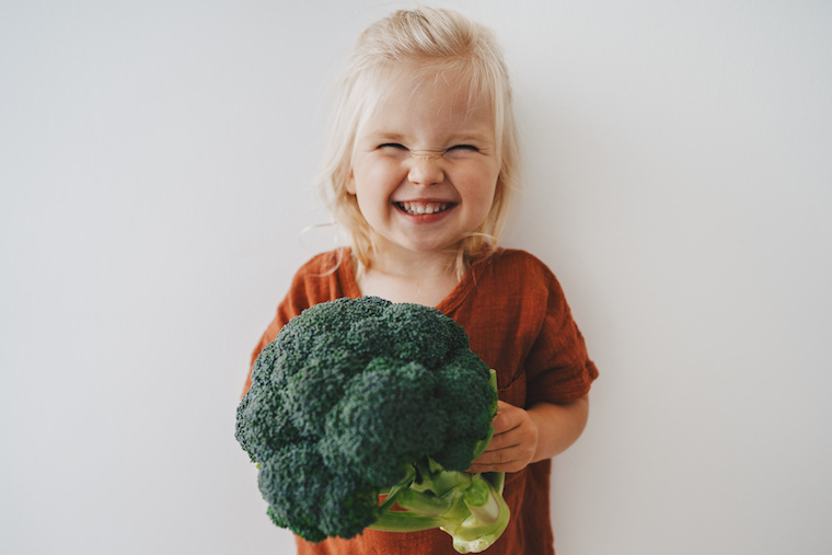 Mädchen hält einen Brokkoli in der Hand und lacht in die Kamera