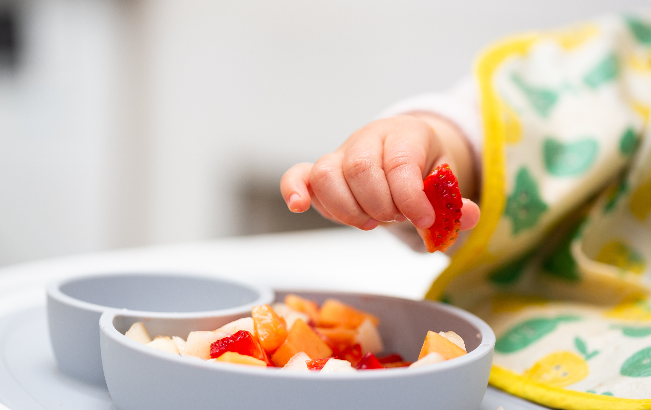 Babyhand greift nach einem Stück Erdbeere von einem Teller voller Obst