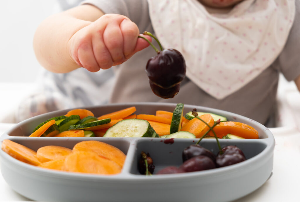 Babyhand greift nach einer Kirsche von einem Teller mit Obst und Gemüse.