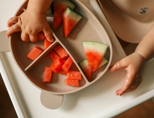 Babyfinger greifen nach einem Stück Wassermelone, das mit mehreren anderen auf einem Teller liegt.