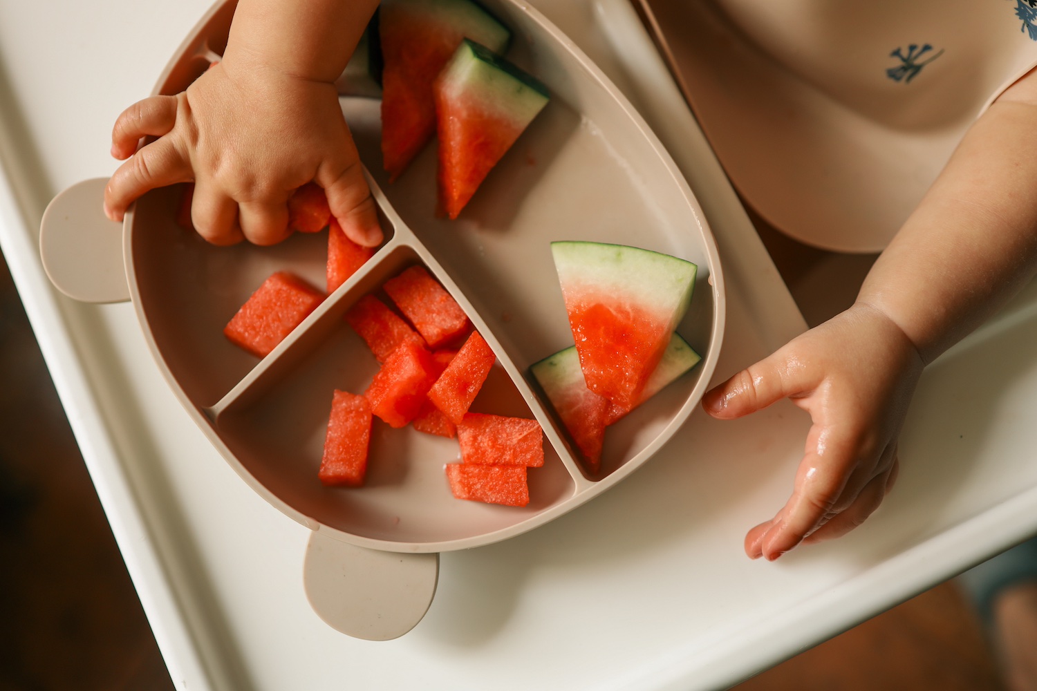 Babyfinger greifen nach einem Stück Wassermelone, das mit mehreren anderen auf einem Teller liegt.