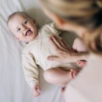 Mutter beugt sich über ein Baby in beigen Body, Vitamin D Gabe ist wichtig für Babys.