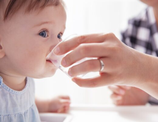 Baby trinkt Wasser aus einem Glas, das ihm seine Mutter hinhält.