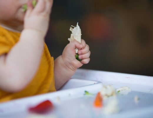 Baby sitzt beim Baby-led Weaning im Hochsitz und die Babyhand greift ein Stück Fleisch, das zusammen mit weiteren Lebensmitteln vor dem Kind liegt.