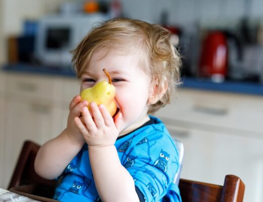 Baby sitzt im Hochstuhl und hält Obst in Form einer Birne in der Hand.